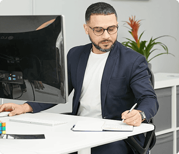 Business professional working at desk with computer and notepad in modern office environment.